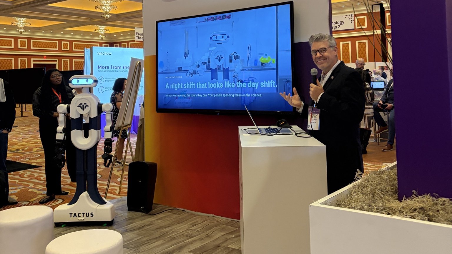 Michael Quick, Tactus AI's Chief Commercial Officer, speaks at the CAP26 Tech Talk Theater beside Elsie by Tactus, the autonomous humanoid lab assistant, under a slide reading "A night shift that looks like the day shift"