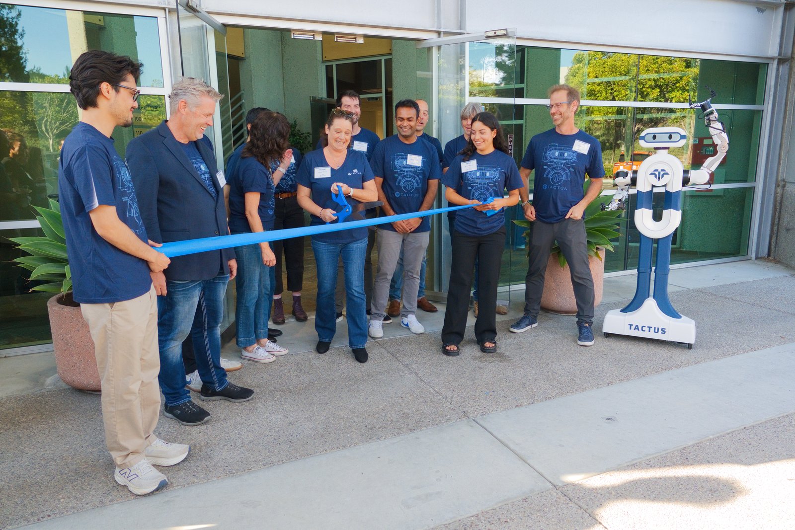 The Tactus AI team cuts a ribbon at the entrance to the San Diego campus, with the humanoid robotic lab assistant beside them