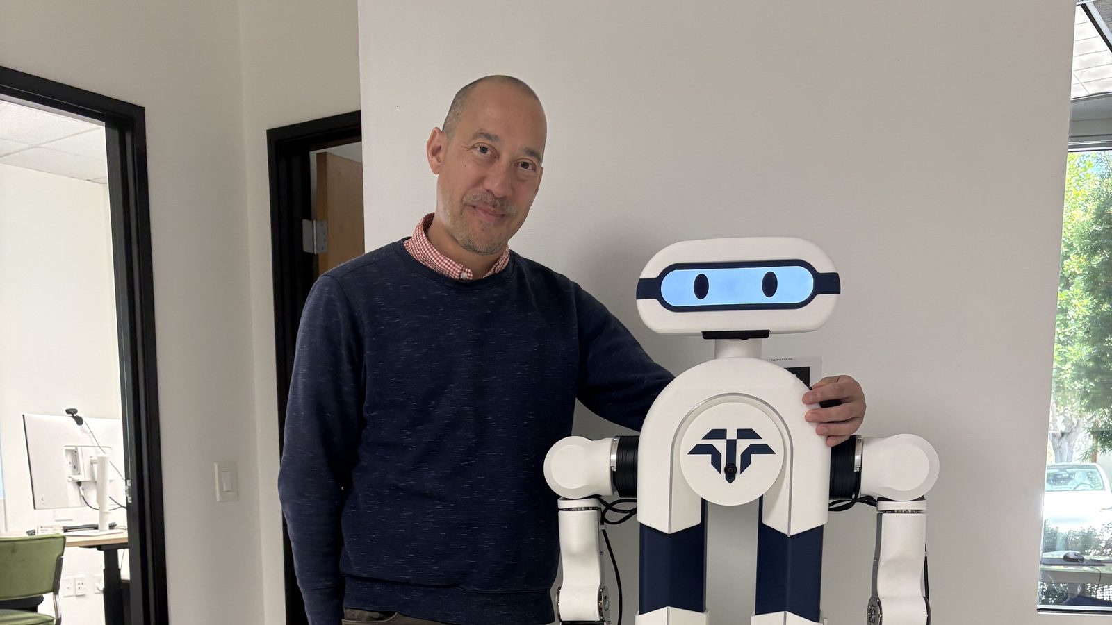 Nathan Wildgrube, Tactus AI's Head of Quality and Regulatory, stands with a hand on the shoulder of Elsie by Tactus, the autonomous humanoid lab assistant, in the Tactus AI office
